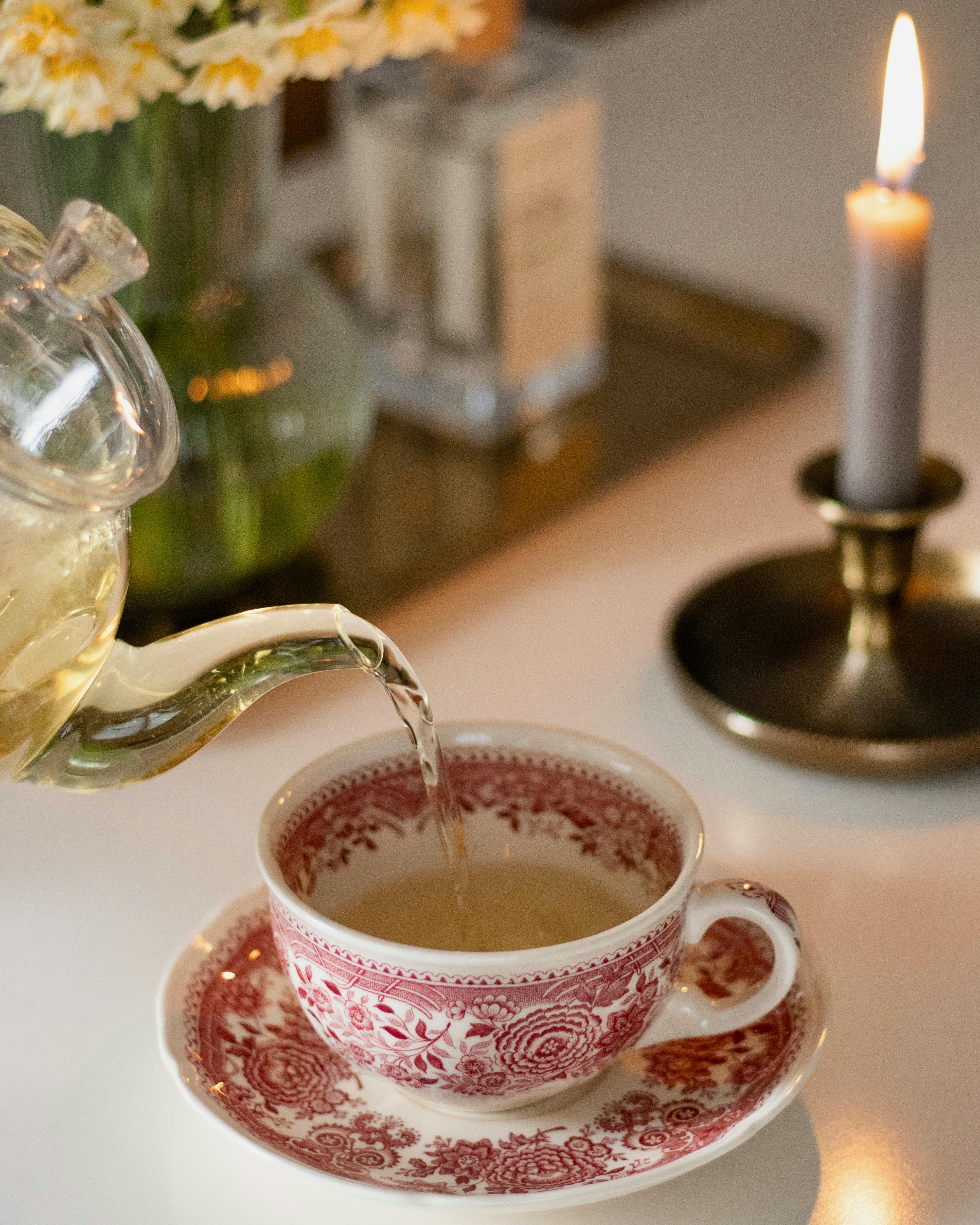 A glass teapot pours tea into an ornate teacup beside a lit candle.