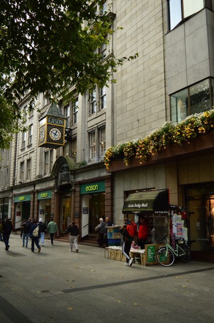 A lively urban street with people walking past stores and a flower-adorned building.