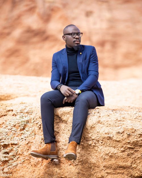 Free Stylish man in a blue suit and eyeglasses sitting on a rock outdoors, exuding elegance. Stock Photo