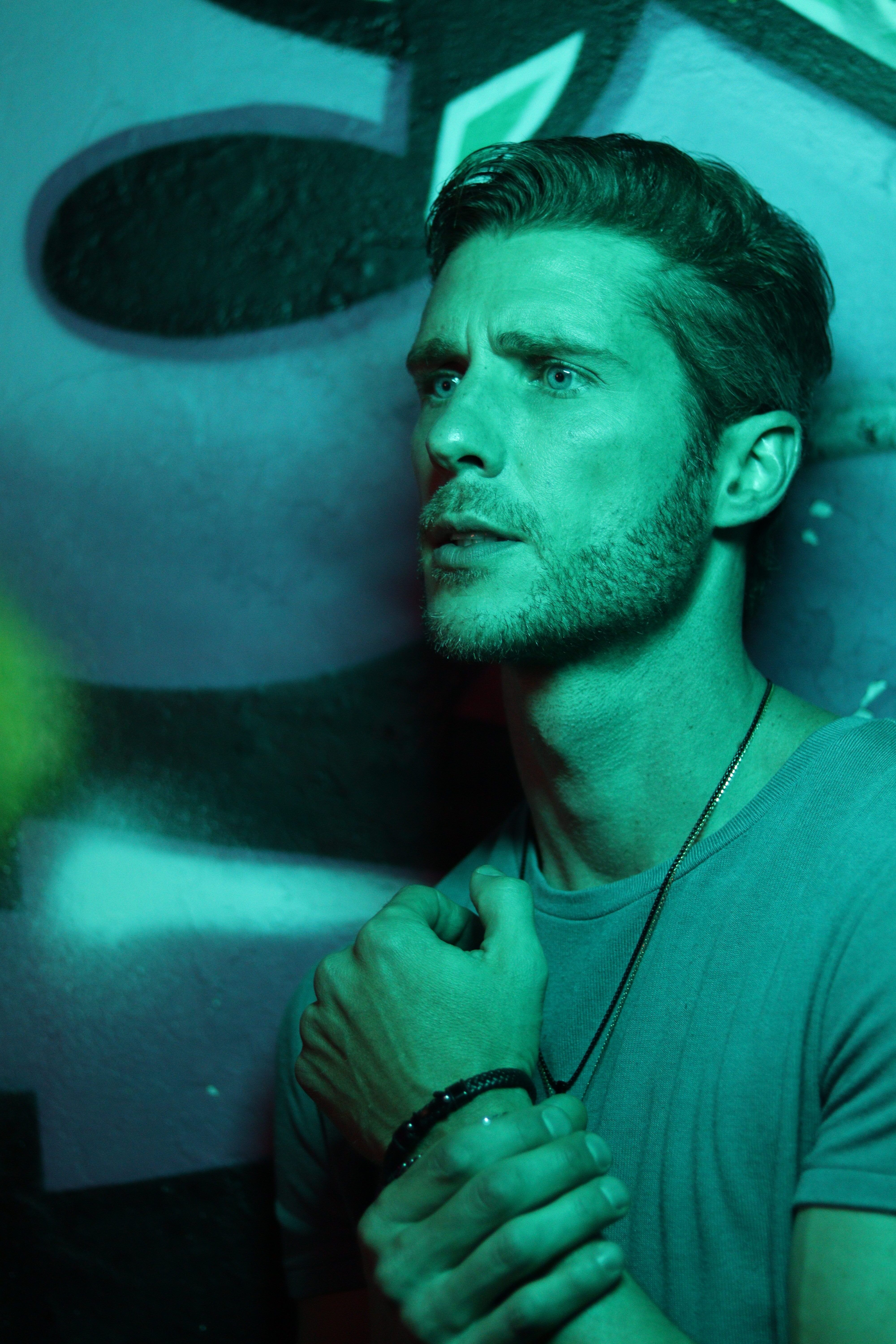 A contemplative man with short hair stands in green light against a graffiti wall, emphasizing mood and depth.