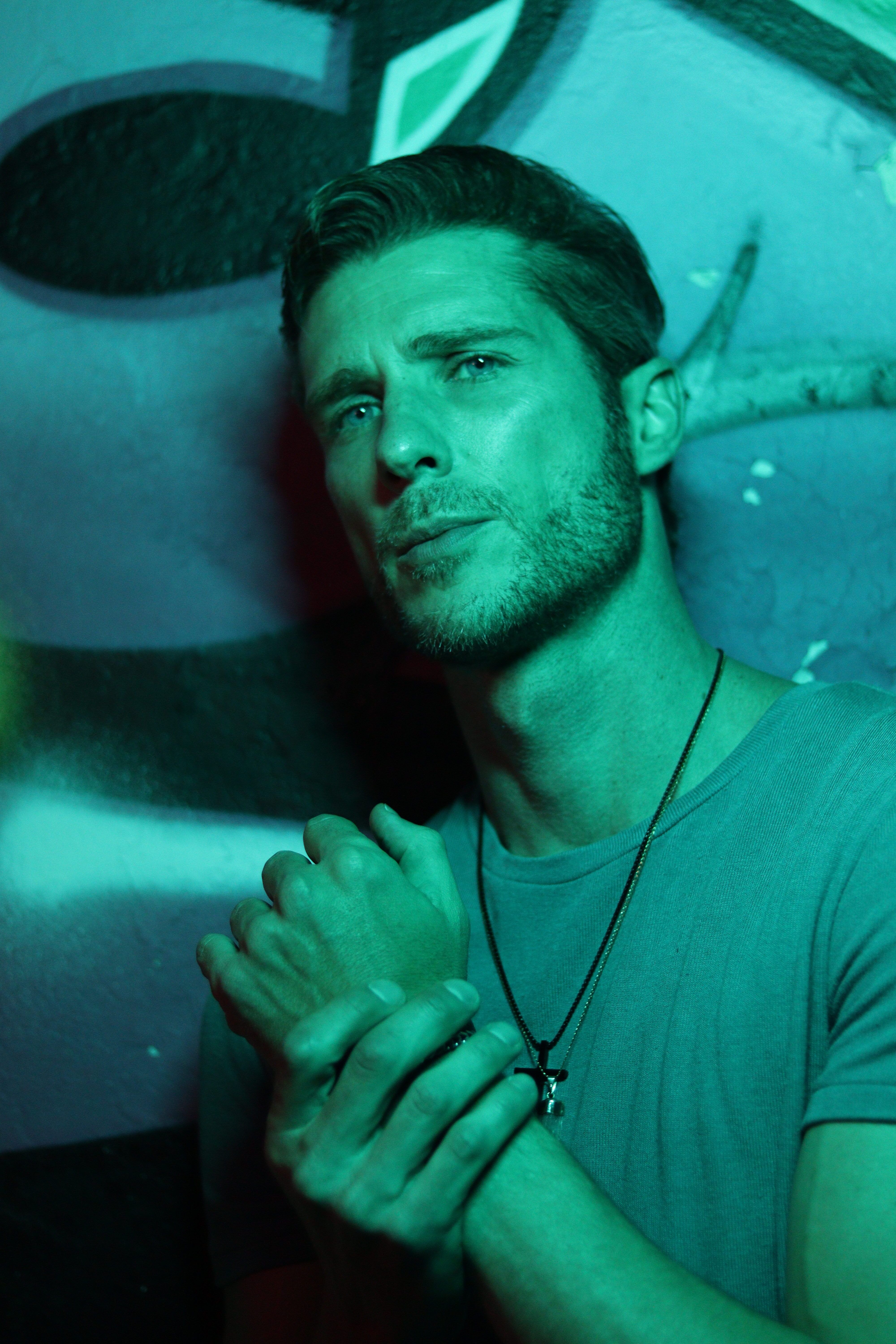 Atmospheric portrait of a man with hands clasped under green lighting, wearing a t-shirt.