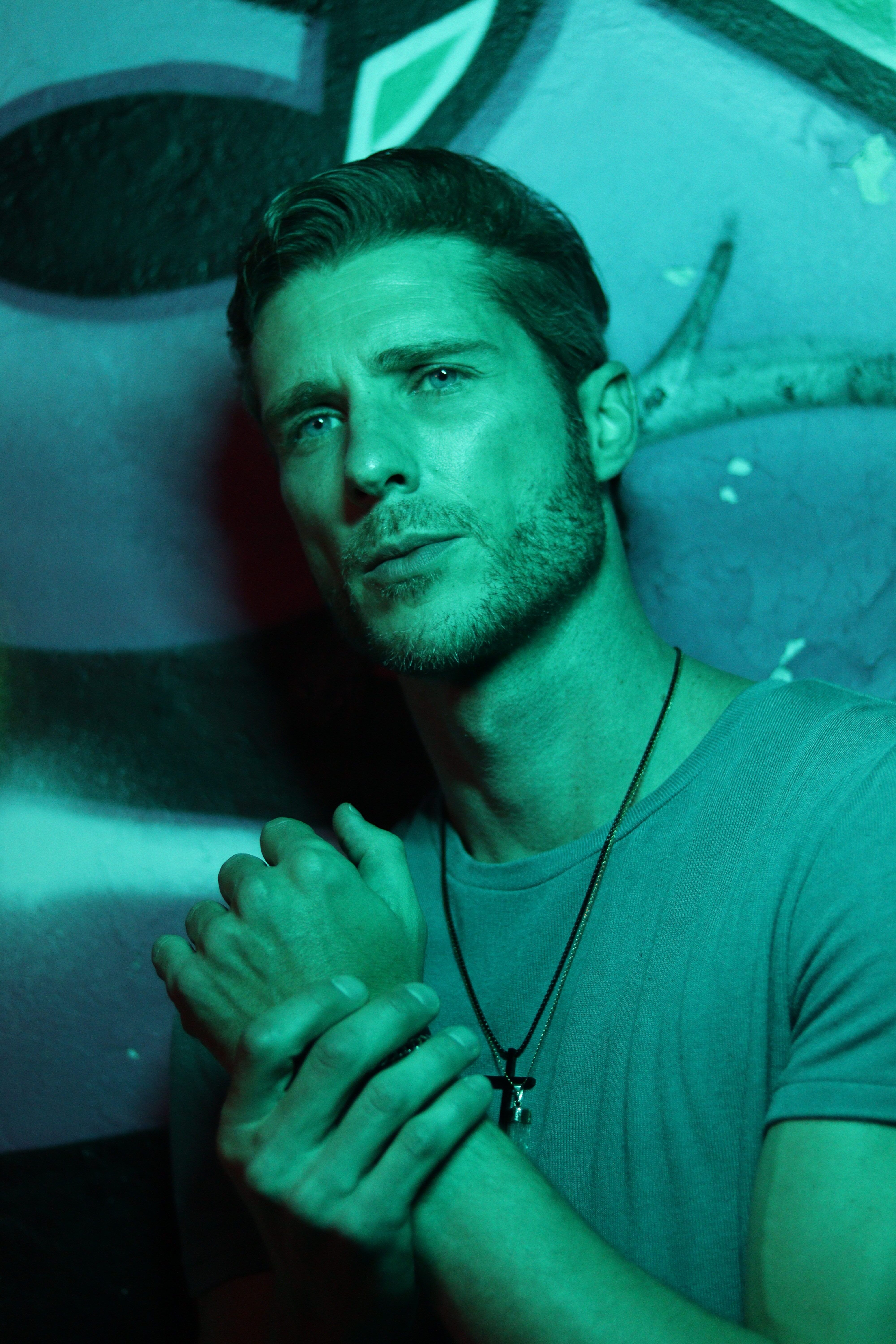 Portrait of a man with short hair holding hands under green lighting, wearing a t-shirt and necklace.