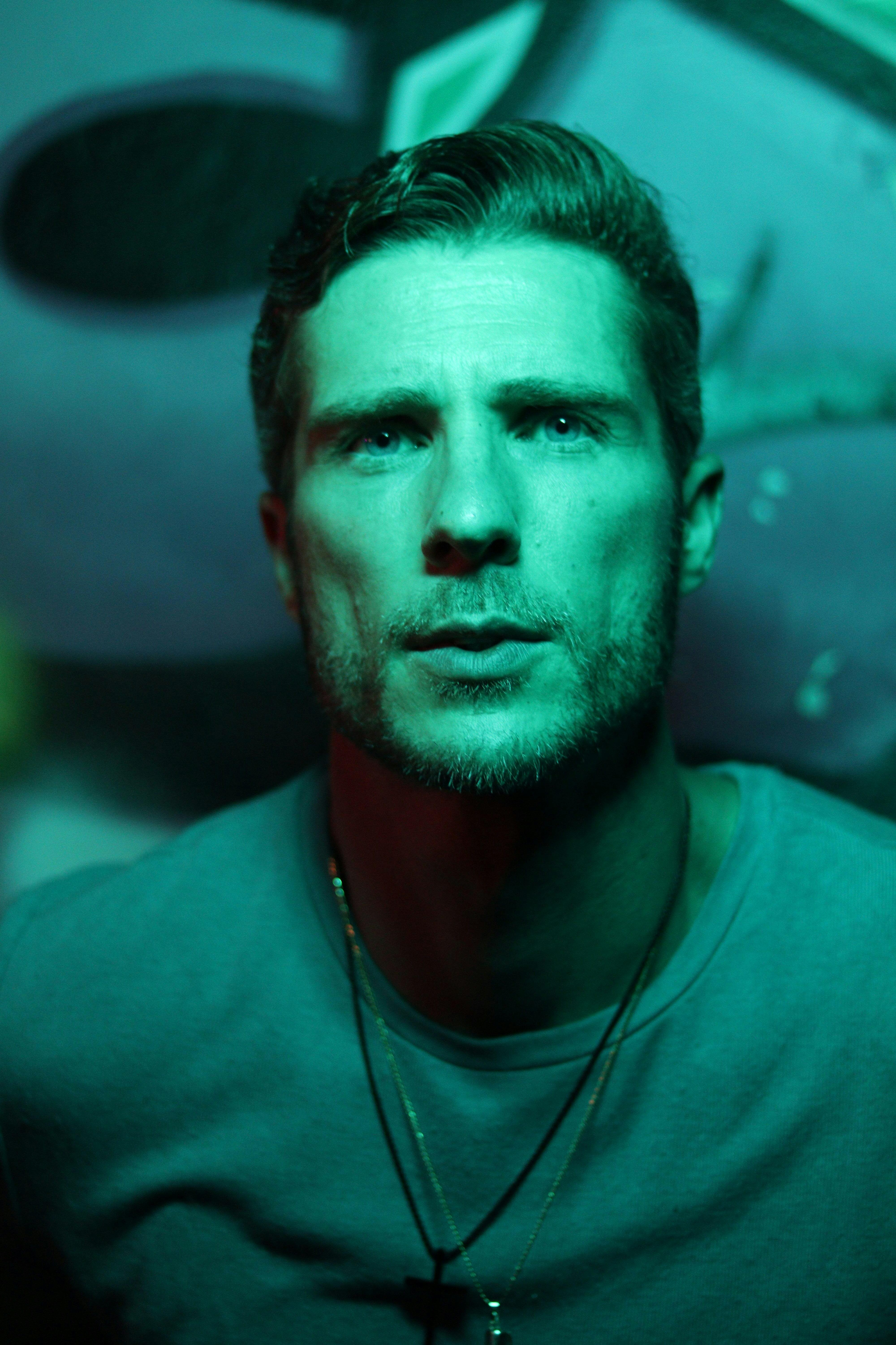 Close-up portrait of a man with dramatic green lighting and necklaces.