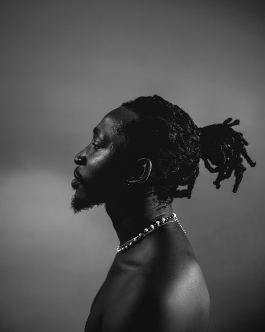 Black and white studio portrait of a shirtless man with braids and a neck chain.