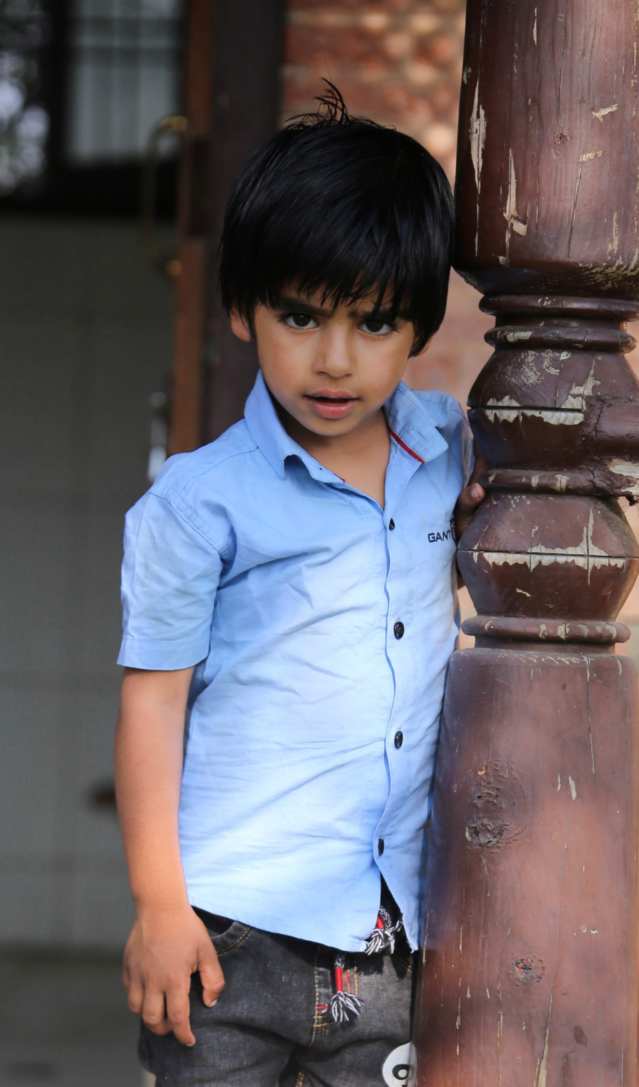 A young boy leans against a rustic wooden column, wearing a blue shirt with a curious expression.