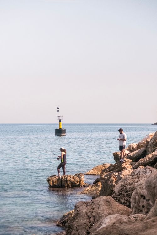 Free Two people fishing on a rocky shoreline near a buoy in the calm sea. Stock Photo