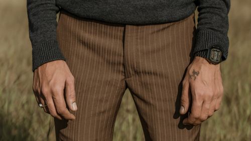 Free Man's hands with a tattoo and wristwatch wearing brown pinstripe trousers in an outdoor setting. Stock Photo