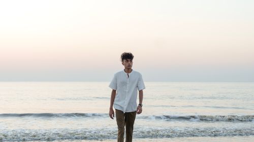 Free Young man with curly hair walking on the beach at sunrise, wearing a white shirt and band collar. Stock Photo