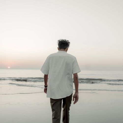 Free A man strolls along the beach at sunset, wearing a white shirt, with the serene sea in the background. Stock Photo
