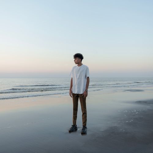 Free A young man in a white shirt stands thoughtfully on a tranquil beach at dusk. Stock Photo