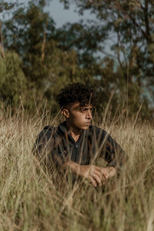 Free A young man with curly hair sits in tall grass, enjoying a peaceful moment outdoors. Stock Photo