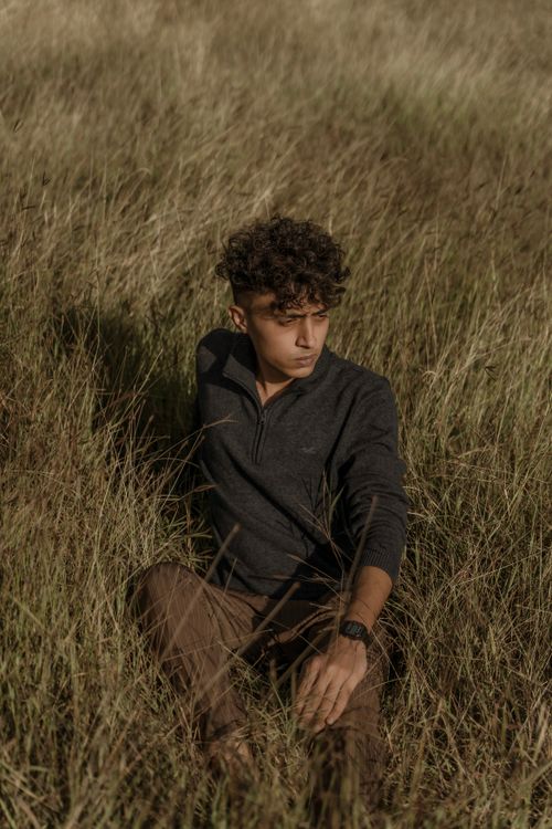 Free A male model with curly hair sits in a grassy field under natural light, conveying relaxed fashion. Stock Photo