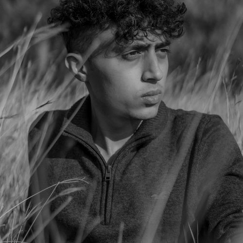 Free Black and white portrait of a man with curly hair sitting in a field, looking contemplative. Stock Photo