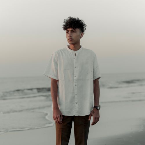 Free Young man standing by a serene beach in a white shirt, showcasing modern fashion. Stock Photo