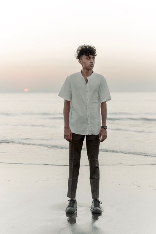Free Young man in white shirt and brown pants stands on beach during sunset, exuding fashion and tranquility. Stock Photo