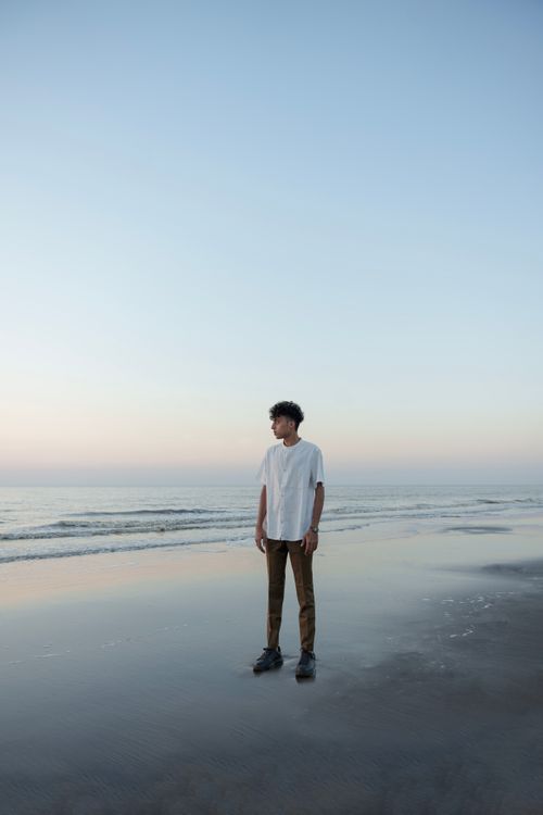 Free A man stands on a tranquil beach at sunrise, wearing casual attire, capturing serenity. Stock Photo