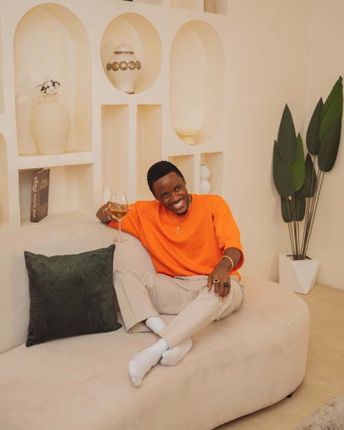 Smiling Man in Orange Sweatshirt and Beige Pants Relaxing with a Glass of Champagne on the Sofa