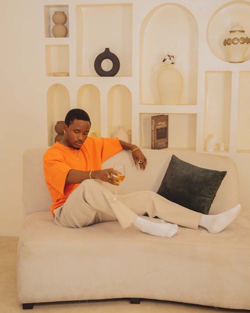 Model in an Orange Sweatshirt with Cropped Sleeves and Beige Pants Sitting on a Sofa with a Glass of Champagne
