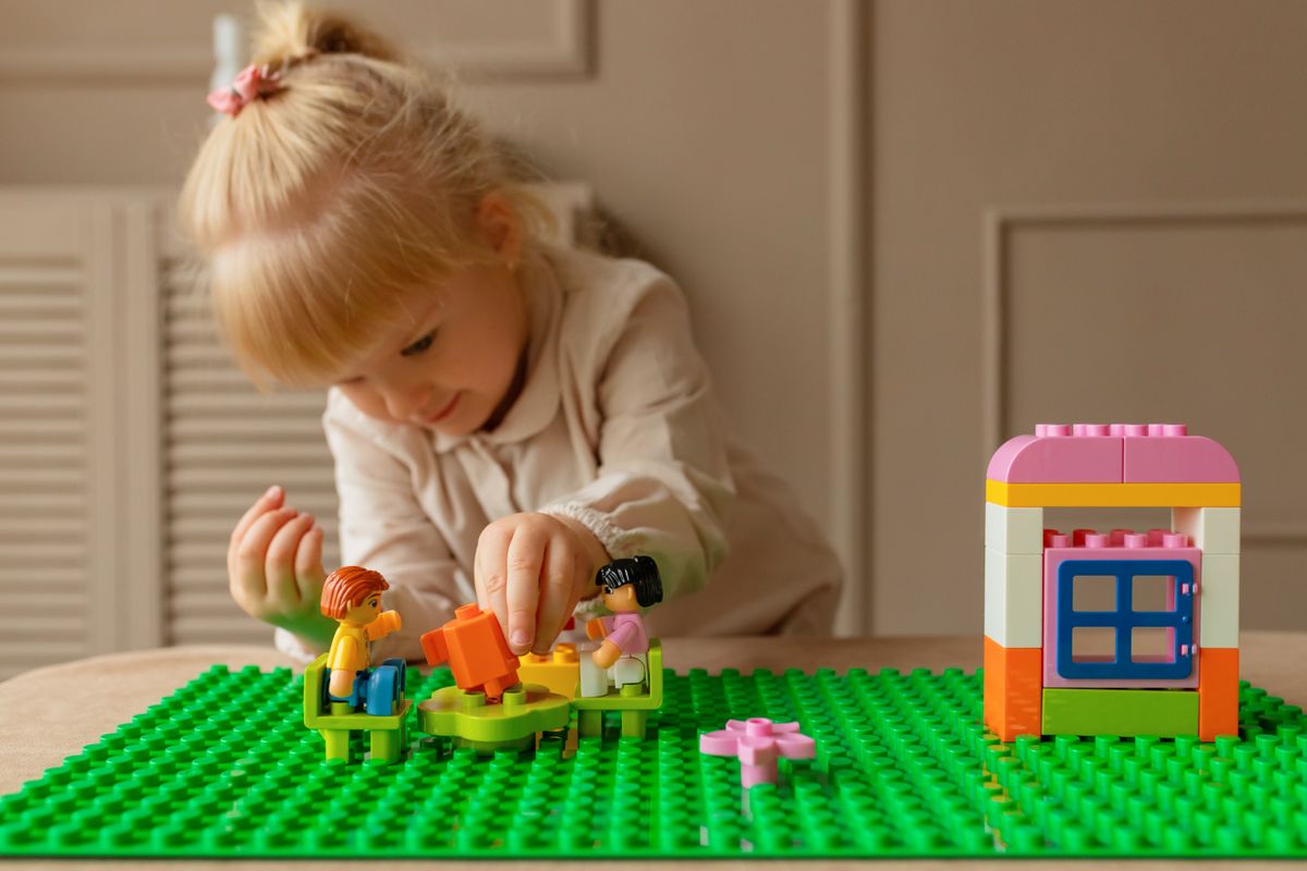 A young girl building with colorful plastic bricks at a table that doubles as LEGO storage space