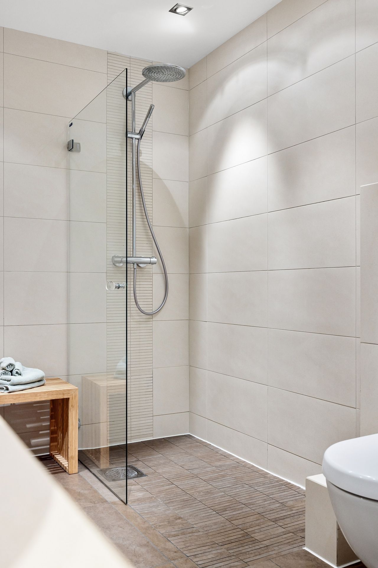Doorless walk-in shower with a frameless fixed glass panel, rainfall showerhead, wood-look tile floor and wooden bench