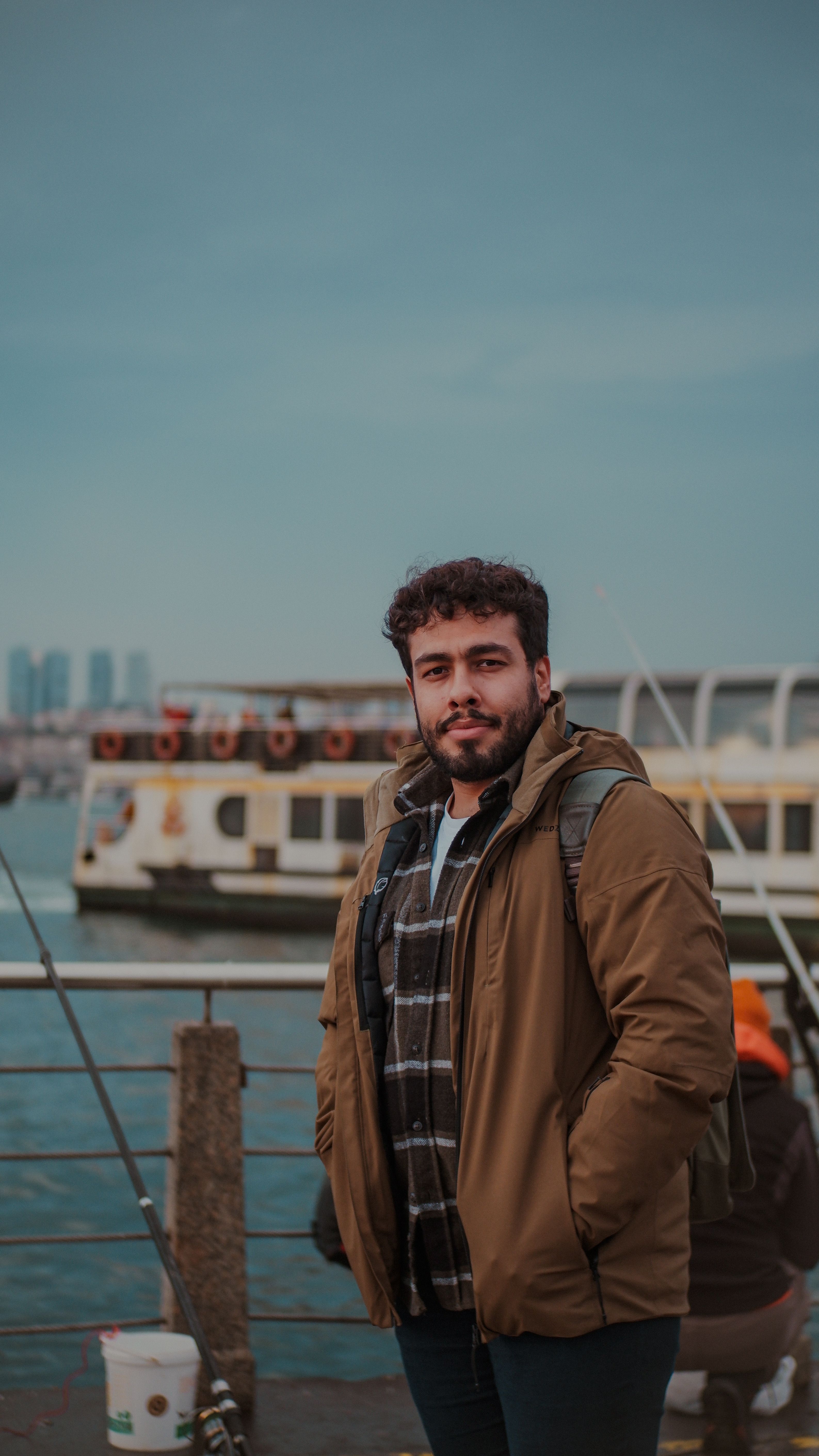 Man wearing a jacket at the Istanbul harbor with a ferry in the background under cloudy skies.