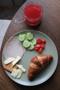 A vibrant breakfast plate with croissant, cheese, ham, cucumbers, and tomatoes, paired with juice.