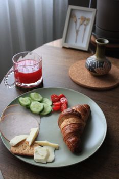 Plate with a croissant, cheese, and fresh veggies on a table with juice.