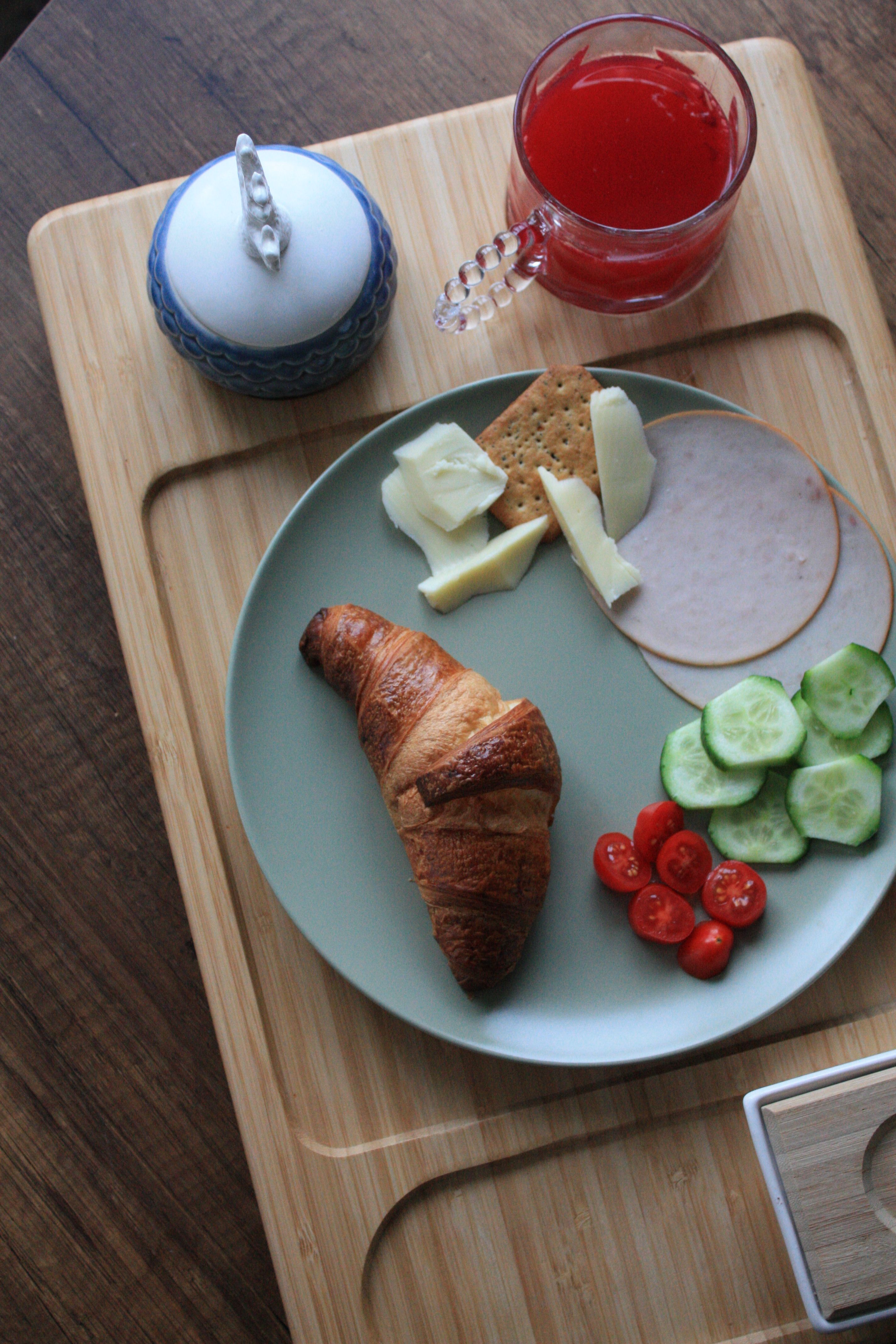 Continental breakfast featuring croissant, cheese, ham, cucumber, and tomatoes on a wooden tray.