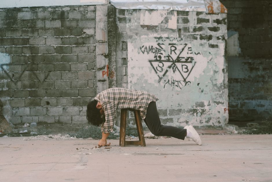A young man in plaid shirt leans over a stool against a graffiti-covered wall.