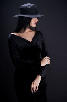 A stylish woman posing in a black dress and hat against a dark background, showcasing elegance.