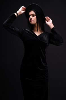 Fashionable brunette woman in black dress and hat posing against dark background.