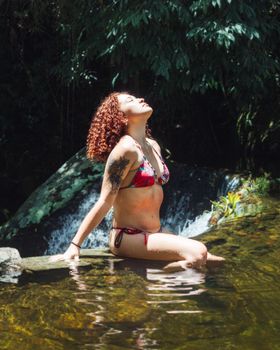 A woman in a bikini relaxes by a waterfall, enjoying the sunny summer day outdoors.