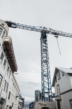 Tower crane in urban setting with buildings under cloudy sky, vertical shot.