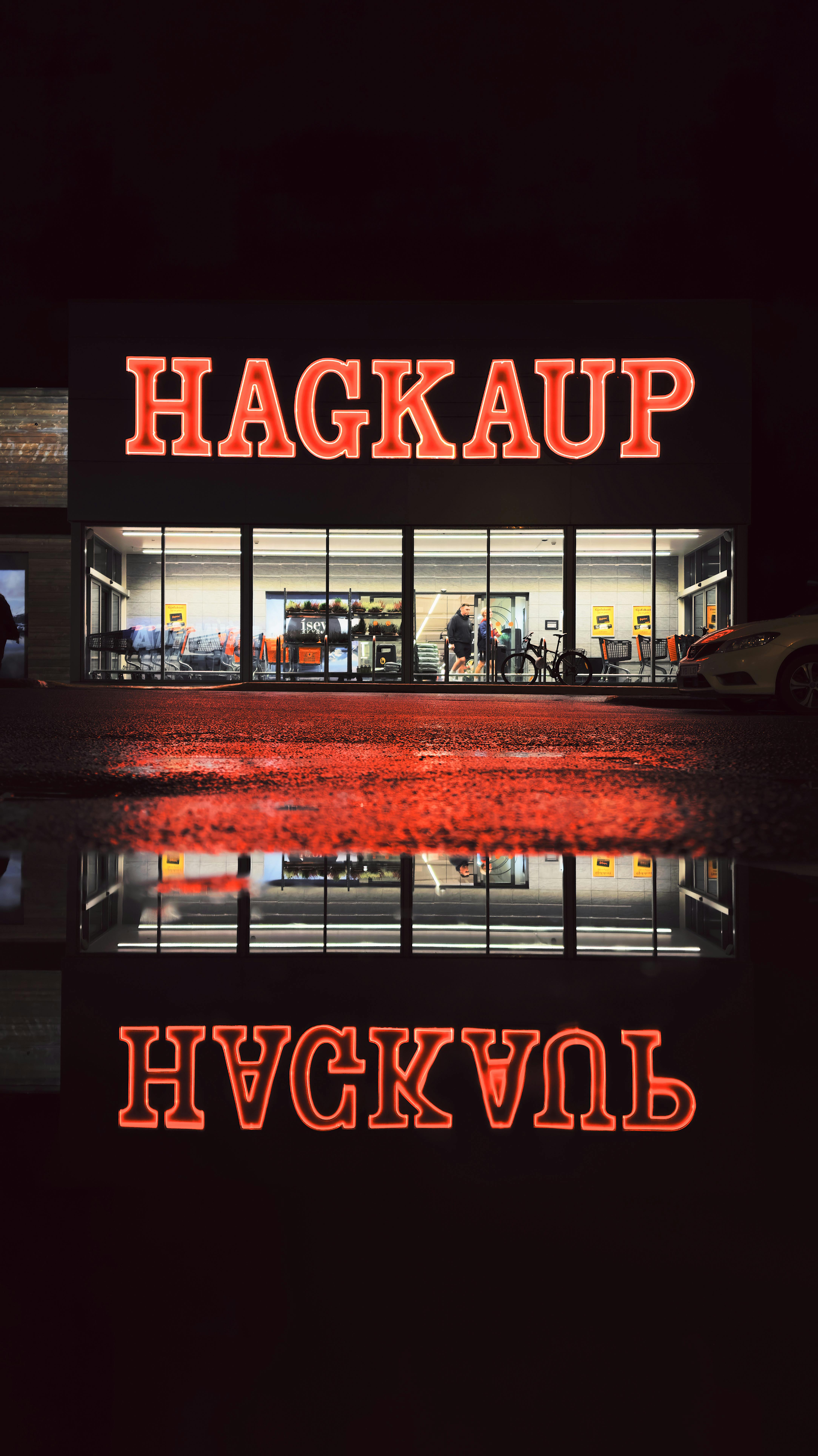 Illuminated Hagkaup store in Reykjavík reflected in a puddle at night.