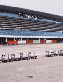 View of Zurich Airport terminal with aligned luggage carts on the tarmac.