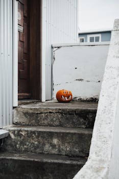A carved pumpkin sits on concrete stairs beside a weathered white wall, evoking a spooky Halloween vibe.