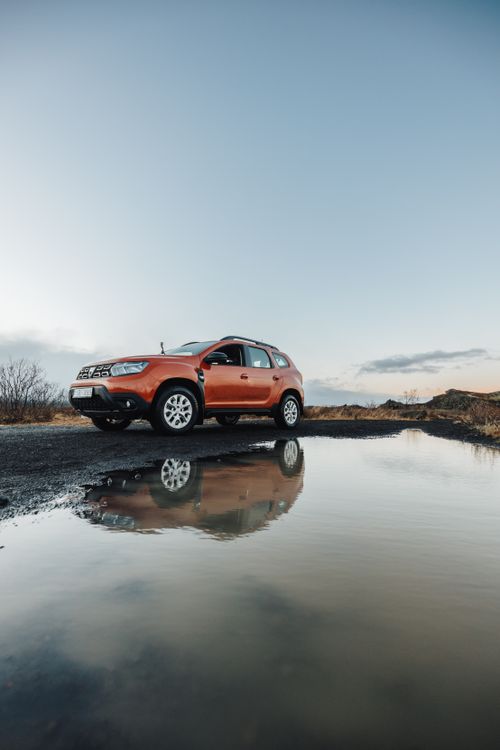 Free An orange SUV parked on a dirt road reflecting in a puddle under a clear sky. Stock Photo