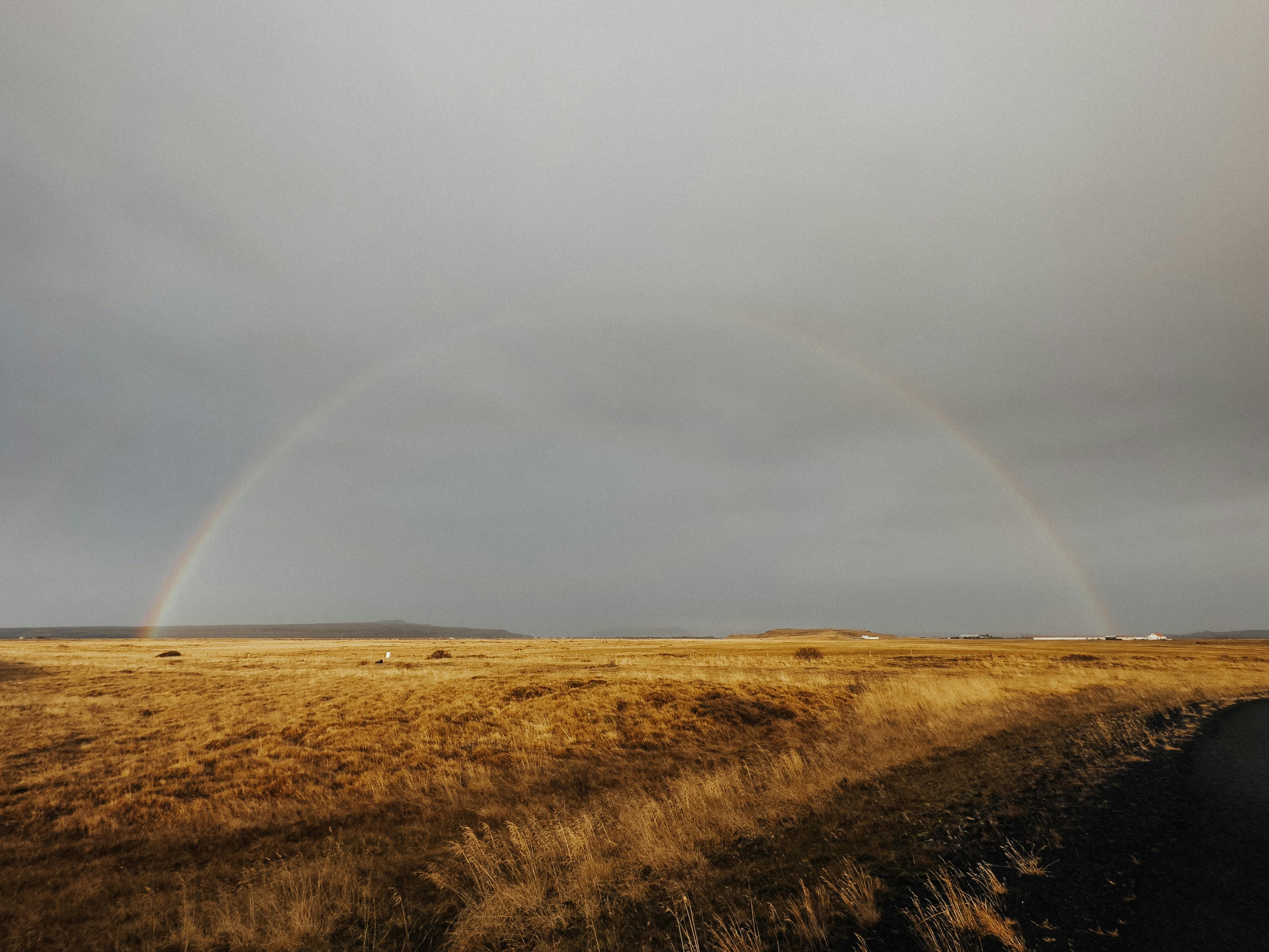 A breathtaking rainbow arcs over the vast grasslands of Flóahreppur, Iceland, under a cloudy sky.