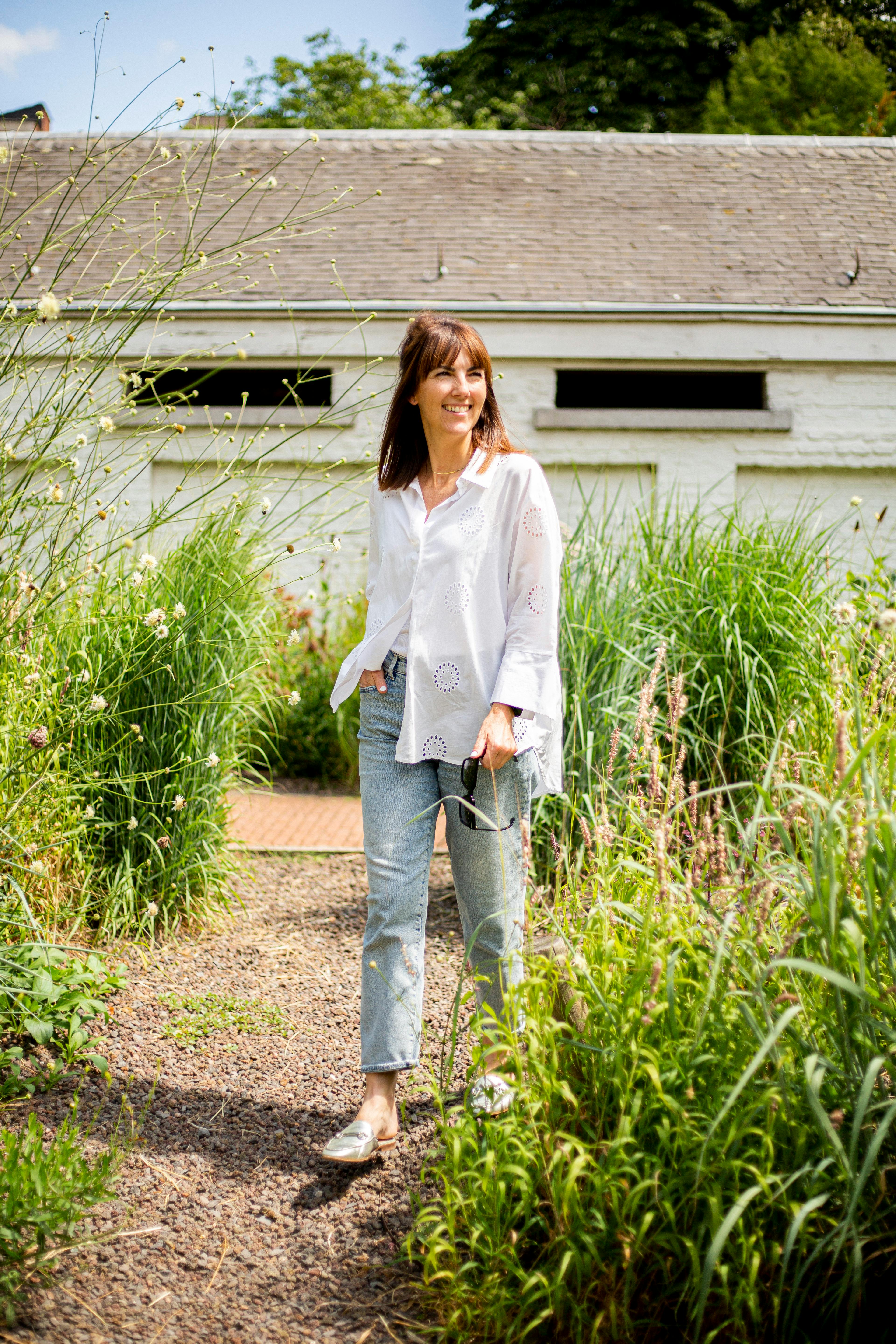 Smiling woman walking along a garden path, surrounded by lush greenery on a sunny day.