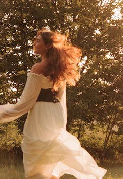 A graceful woman with flowing hair dances outdoors in a white dress, backlit by the sun.