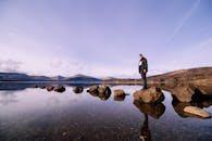 一名男子站在宁静湖泊的岩石上，山脉倒映在晴朗的天空下