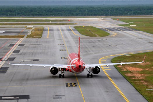 A red airplane taxis on an empty airport runway with surrounding greenery.