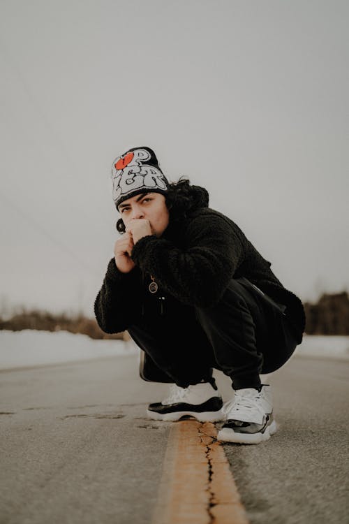 Man in Hat and Black Clothes Squatting on Road