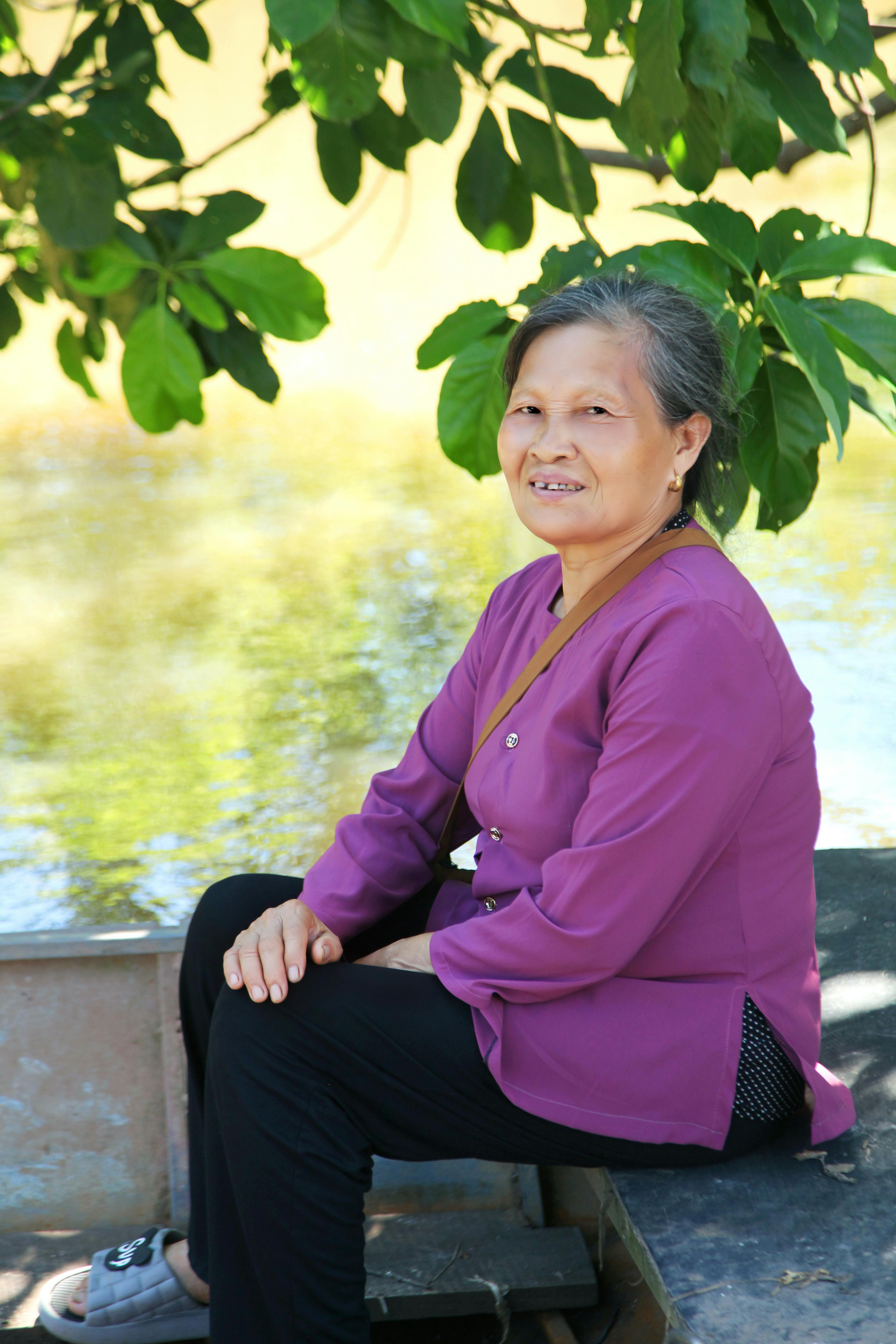 Portrait of an elderly Asian woman sitting outdoors by a lakeshore in a stylish purple shirt.