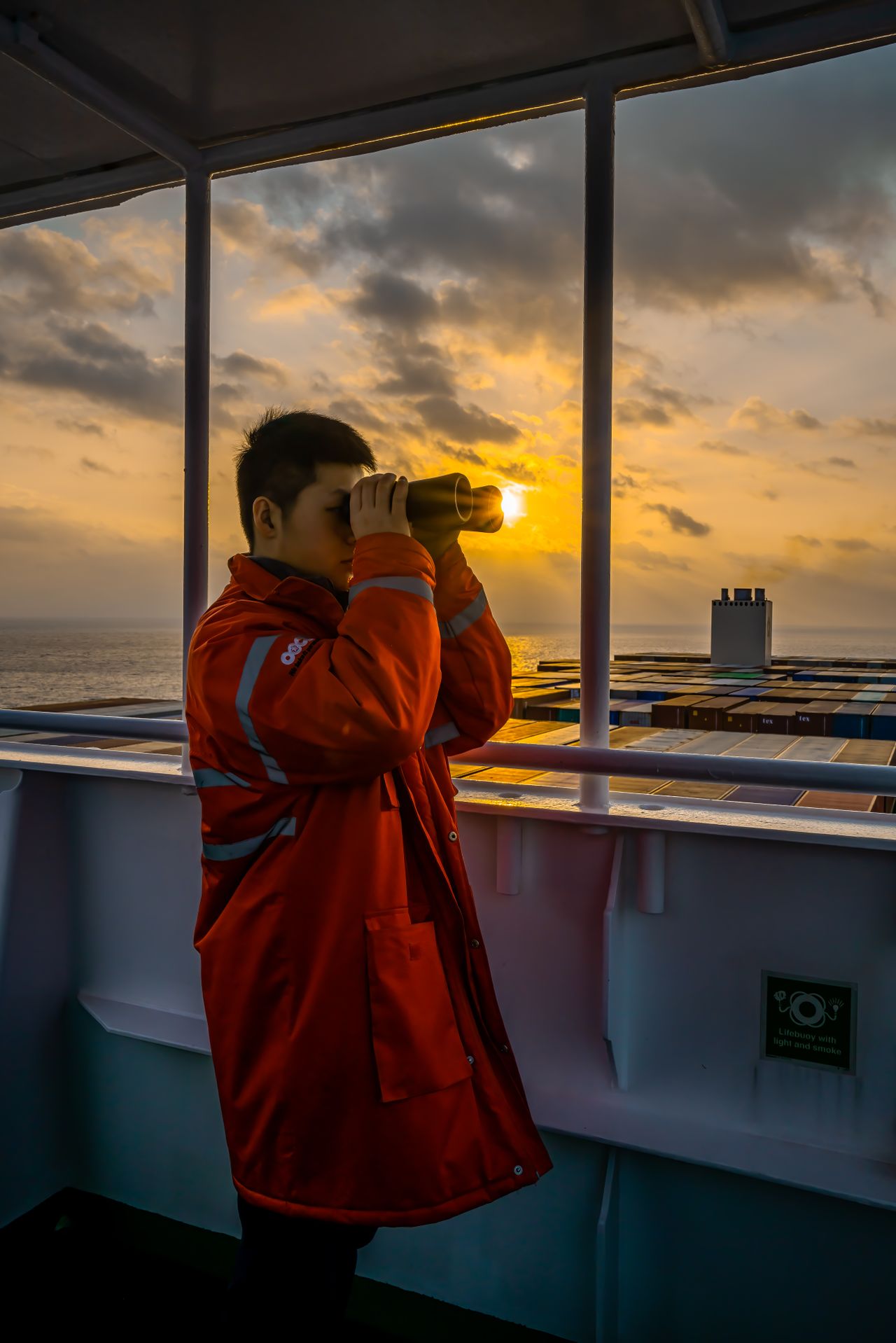 Man in red jacket using binoculars on a ship at sunset, working at sea.