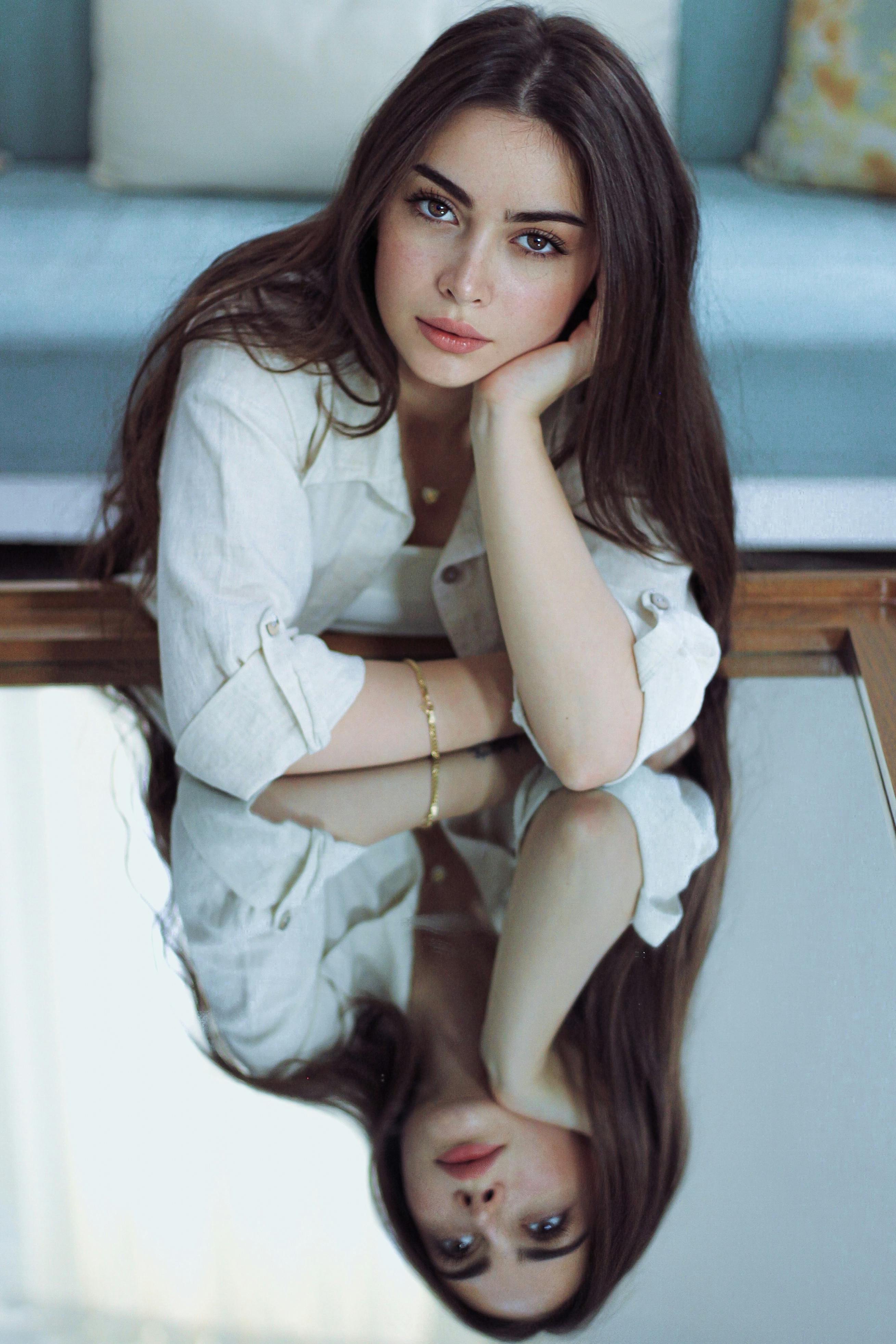 Portrait of a woman with brown hair in a white shirt, her reflection visible on a glass table.