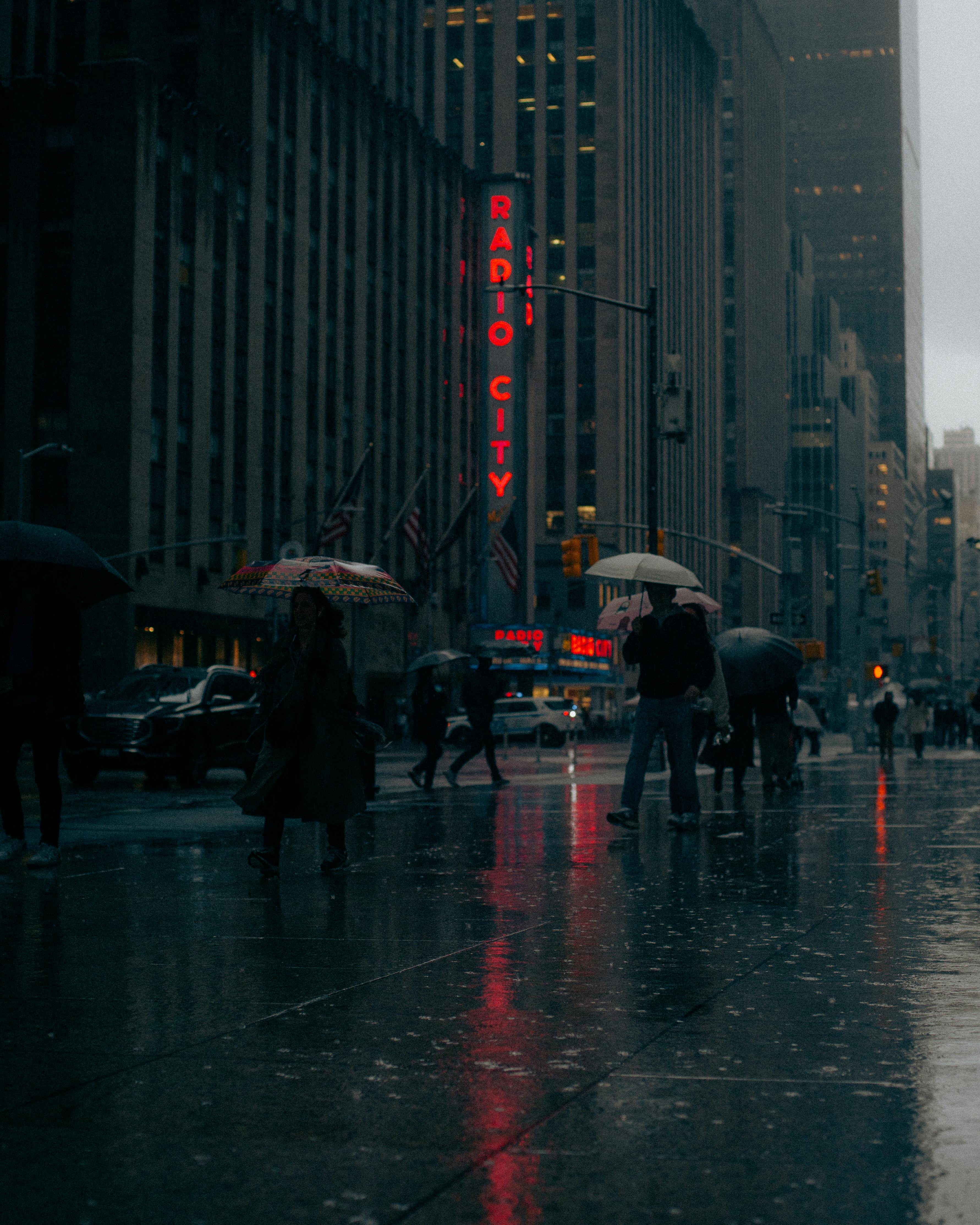 People walking with umbrellas in rain near Radio City, New York City at night.