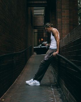 A man in a tank top leans against a brick wall, engrossed in his smartphone in an urban alleyway.