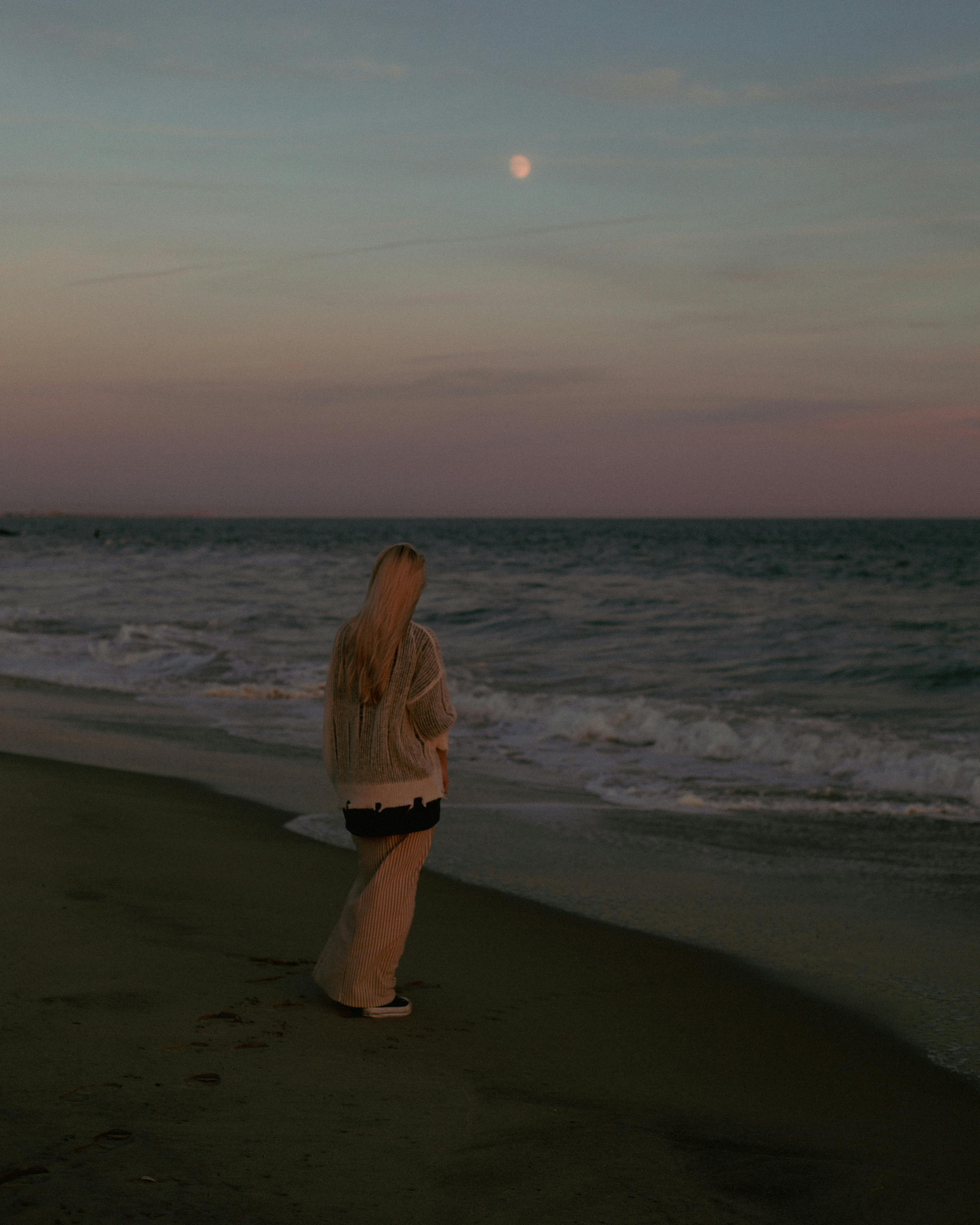 A solitary woman stands on a serene beach, gazing at the ocean and moon at dusk.