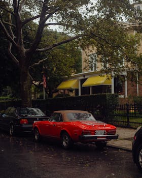 A red vintage convertible car parked on a rainy city street with trees and classic houses.